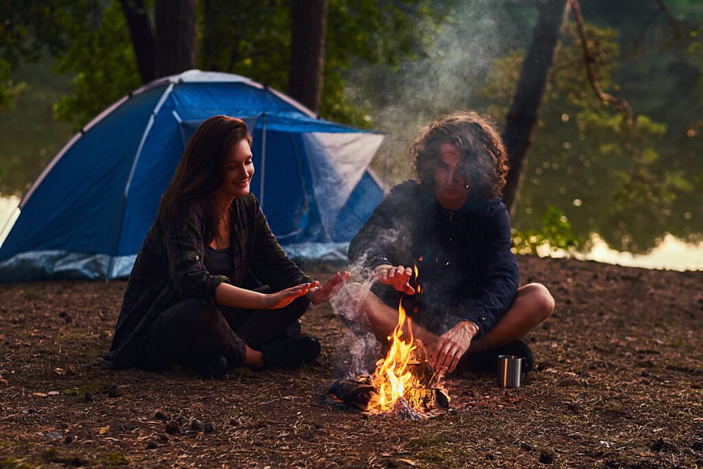 Junges Pärchen sitzt im Wald vor einem Lagerfeuer mit Camping-Zelten im Hintergrund.